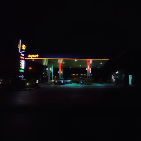A petrol station is illuminated at night, featuring a bright canopy with red and yellow branding. Several cars are parked under the shelter, and the surrounding area is enveloped in darkness, highlighting the contrast with the well-lit petrol station.
