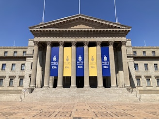 A large neoclassical building with tall columns at the entrance. There are three banners hanging between the columns, with two blue banners on the sides and a yellow one in the middle. The banners display the WITS University logo and commemorate its 100th anniversary. The exterior is symmetrical, with multiple windows and a grand staircase leading up to the entrance.