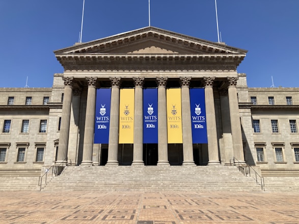 A large neoclassical building with tall columns at the entrance. There are three banners hanging between the columns, with two blue banners on the sides and a yellow one in the middle. The banners display the WITS University logo and commemorate its 100th anniversary. The exterior is symmetrical, with multiple windows and a grand staircase leading up to the entrance.