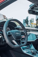 The image captures the interior of a modern car, featuring a prominently displayed steering wheel with the Audi logo. The dashboard and console are sleek, with digital displays and touch controls. Natural light streams through the windshield, illuminating the high-quality materials and design features within the vehicle.