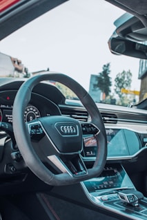 The image captures the interior of a modern car, featuring a prominently displayed steering wheel with the Audi logo. The dashboard and console are sleek, with digital displays and touch controls. Natural light streams through the windshield, illuminating the high-quality materials and design features within the vehicle.