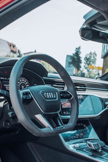 The image captures the interior of a modern car, featuring a prominently displayed steering wheel with the Audi logo. The dashboard and console are sleek, with digital displays and touch controls. Natural light streams through the windshield, illuminating the high-quality materials and design features within the vehicle.
