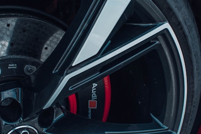 A close-up view of a car wheel and brake disc, including details like text that reads 'Audi' and 'Made in Germany.' The wheel features a black and metallic silver design, while the brake caliper is red.