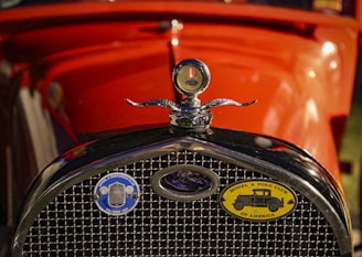 The front view of a vintage car featuring a chrome radiator grille and an emblem with a detailed design. Two badges are visible on the grille, including one for the Model A Ford Club of America. The car is painted in a vibrant orange color, accented by the shiny metallic elements.