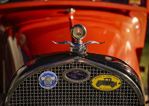 The front view of a vintage car featuring a chrome radiator grille and an emblem with a detailed design. Two badges are visible on the grille, including one for the Model A Ford Club of America. The car is painted in a vibrant orange color, accented by the shiny metallic elements.