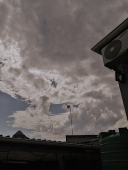 A cloudy sky with a break showing blue sky. In the foreground, there is a rooftop, a green water tank, and a pole with a security camera. An air conditioning unit is mounted on the wall.