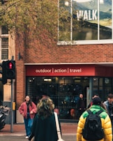 A busy street scene with people walking past a store that promotes outdoor, action, and travel activities. The store has a brick exterior and large windows displaying advertisements for walking. Pedestrians are wearing jackets, one with a bright yellow and green coat, another with a pink jacket, and several people in casual attire.