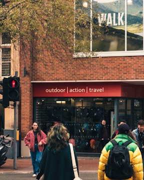 A busy street scene with people walking past a store that promotes outdoor, action, and travel activities. The store has a brick exterior and large windows displaying advertisements for walking. Pedestrians are wearing jackets, one with a bright yellow and green coat, another with a pink jacket, and several people in casual attire.