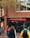 A busy street scene with people walking past a store that promotes outdoor, action, and travel activities. The store has a brick exterior and large windows displaying advertisements for walking. Pedestrians are wearing jackets, one with a bright yellow and green coat, another with a pink jacket, and several people in casual attire.