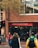 A busy street scene with people walking past a store that promotes outdoor, action, and travel activities. The store has a brick exterior and large windows displaying advertisements for walking. Pedestrians are wearing jackets, one with a bright yellow and green coat, another with a pink jacket, and several people in casual attire.