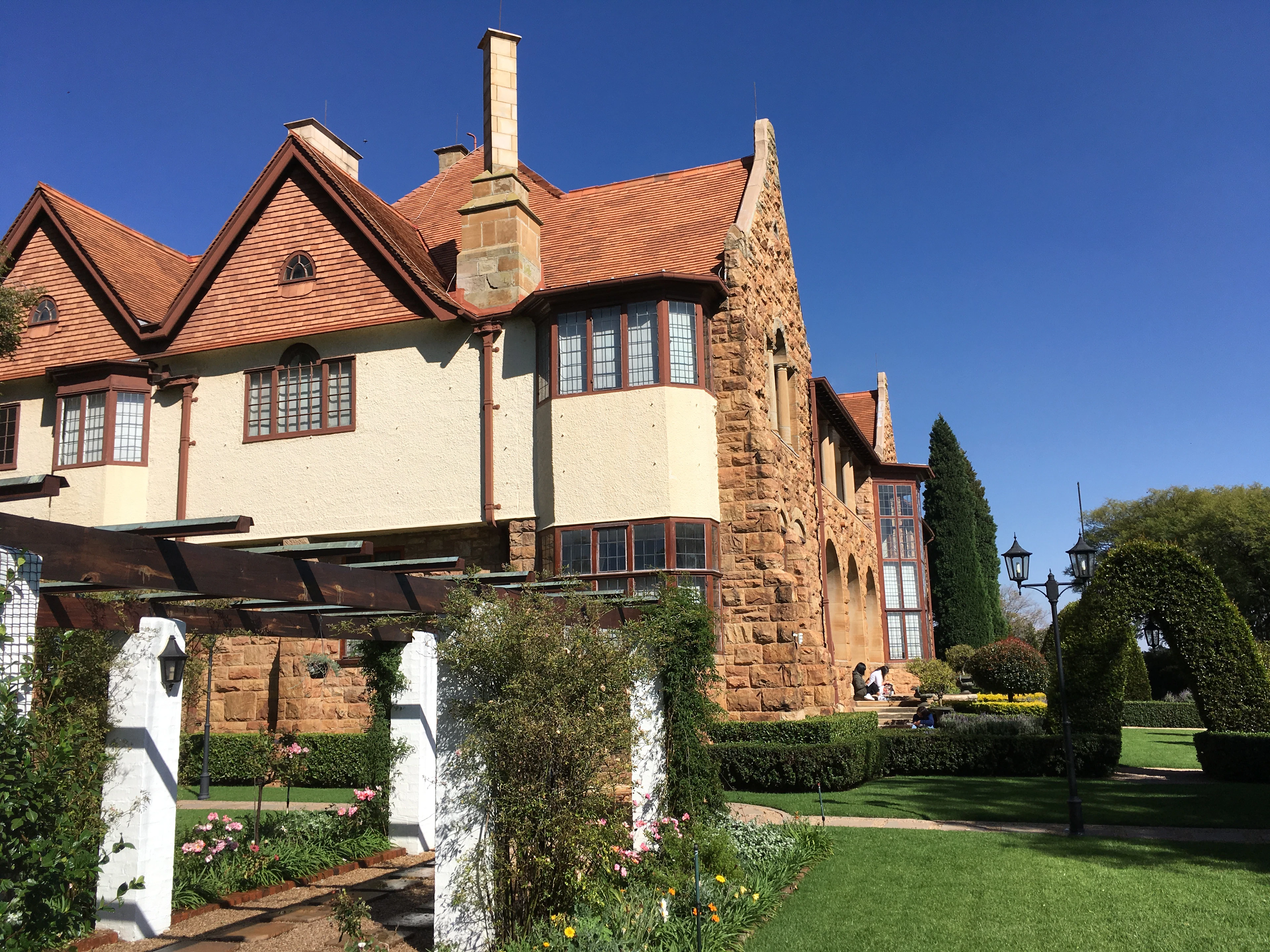 A grand sandstone and cream gabled manor house with a deep red-tiled roof in Parktown, Johannesburg, seen across a formal rose garden with a white pergola and clipped hedges under a deep blue sky.