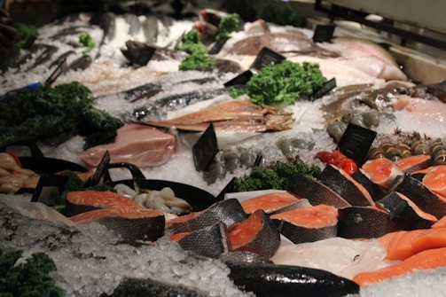 A fresh seafood display on a bed of ice featuring various types of fish and shellfish. Salmon fillets are prominently placed alongside other cuts of fish. Green garnishes and small labels are scattered throughout the display.