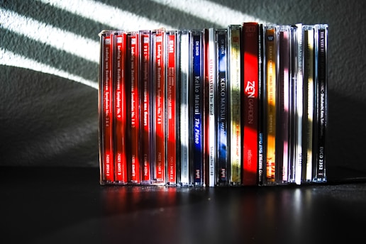 A collection of music CDs is arranged side by side on a black surface. The CDs have various colorful spine labels, with many featuring red, blue, white, and a mix of other hues. The background is a textured gray wall with light streaming through blinds, creating diagonal shadow patterns on the wall and partially on the CDs.