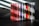 A collection of music CDs is arranged side by side on a black surface. The CDs have various colorful spine labels, with many featuring red, blue, white, and a mix of other hues. The background is a textured gray wall with light streaming through blinds, creating diagonal shadow patterns on the wall and partially on the CDs.