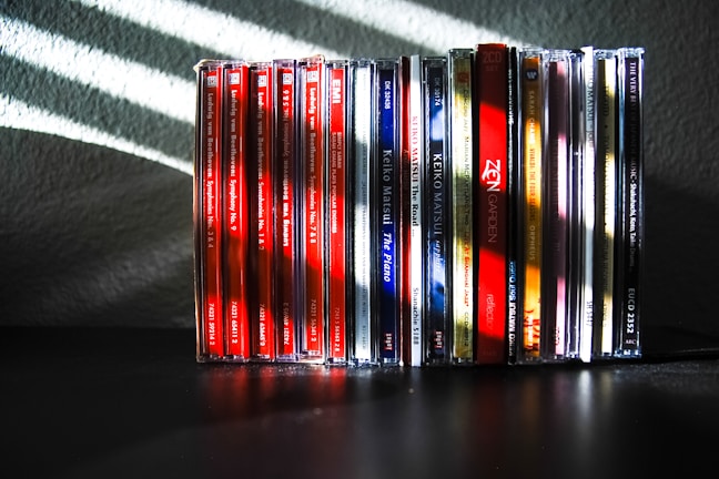 A collection of music CDs is arranged side by side on a black surface. The CDs have various colorful spine labels, with many featuring red, blue, white, and a mix of other hues. The background is a textured gray wall with light streaming through blinds, creating diagonal shadow patterns on the wall and partially on the CDs.