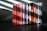 A collection of music CDs is arranged side by side on a black surface. The CDs have various colorful spine labels, with many featuring red, blue, white, and a mix of other hues. The background is a textured gray wall with light streaming through blinds, creating diagonal shadow patterns on the wall and partially on the CDs.