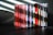 A collection of music CDs is arranged side by side on a black surface. The CDs have various colorful spine labels, with many featuring red, blue, white, and a mix of other hues. The background is a textured gray wall with light streaming through blinds, creating diagonal shadow patterns on the wall and partially on the CDs.