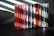 A collection of music CDs is arranged side by side on a black surface. The CDs have various colorful spine labels, with many featuring red, blue, white, and a mix of other hues. The background is a textured gray wall with light streaming through blinds, creating diagonal shadow patterns on the wall and partially on the CDs.