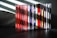 A collection of music CDs is arranged side by side on a black surface. The CDs have various colorful spine labels, with many featuring red, blue, white, and a mix of other hues. The background is a textured gray wall with light streaming through blinds, creating diagonal shadow patterns on the wall and partially on the CDs.
