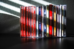 A collection of music CDs is arranged side by side on a black surface. The CDs have various colorful spine labels, with many featuring red, blue, white, and a mix of other hues. The background is a textured gray wall with light streaming through blinds, creating diagonal shadow patterns on the wall and partially on the CDs.