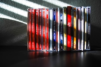 A collection of music CDs is arranged side by side on a black surface. The CDs have various colorful spine labels, with many featuring red, blue, white, and a mix of other hues. The background is a textured gray wall with light streaming through blinds, creating diagonal shadow patterns on the wall and partially on the CDs.