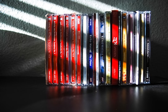 A collection of music CDs is arranged side by side on a black surface. The CDs have various colorful spine labels, with many featuring red, blue, white, and a mix of other hues. The background is a textured gray wall with light streaming through blinds, creating diagonal shadow patterns on the wall and partially on the CDs.