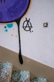 Graffiti on a wall features a large purple circle with a dripping black line and an anarchist symbol next to an exposed light switch. The steps below are covered with shattered glass.