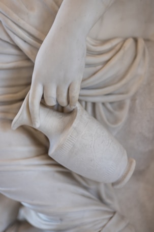 A detailed view of a marble sculpture showing a hand delicately holding an intricately carved jug. The flowing drapery of the sculpture creates a sense of fluidity and grace.