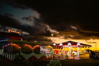 A vibrant carnival scene with a colorful carousel illuminated by bright lights stands under a dramatic, cloudy sky at dusk. The word 'MUNDIAL' is seen lit up in the background. There's a sense of activity, with the brightly lit attractions contrasting against the dark clouds.