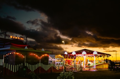 A vibrant carnival scene with a colorful carousel illuminated by bright lights stands under a dramatic, cloudy sky at dusk. The word 'MUNDIAL' is seen lit up in the background. There's a sense of activity, with the brightly lit attractions contrasting against the dark clouds.