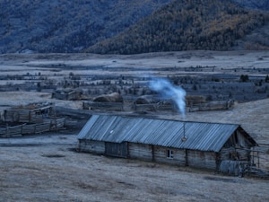 A rustic wooden building with a steeply pitched corrugated roof is set in an open field. Smoke is rising from a chimney, indicating activity inside. The landscape includes several fenced-off circular haystacks, and a dense forest covering the mountains in the background.