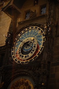 An intricate astronomical clock with detailed golden Roman numerals and zodiac symbols on a blue and red background. The clock is adorned with sculptural elements and figures on its sides, set against an ornate architectural structure.