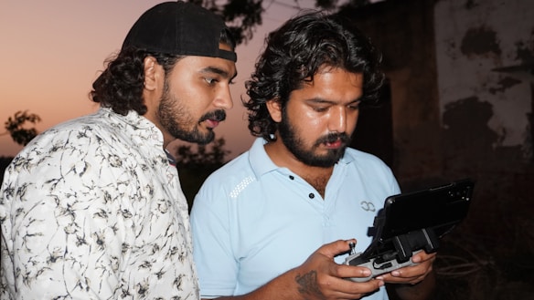 Two men are focused on operating a drone controller with a monitor attached. The person on the left is wearing a cap and a patterned shirt, while the person on the right is wearing a light blue polo shirt. They appear to be outdoors during sunset, with silhouetted trees and a building wall in the background.