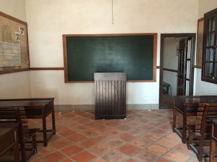 A classroom with a chalkboard on the front wall surrounded by a wooden frame. There are wooden desks and chairs arranged facing the chalkboard. The room has tiled floors and slightly textured beige walls. On the left wall, there is a large board with some documents or posters pinned to it, and on the right, an open doorway leading to another room is visible. A large window is present on the right side of the room.