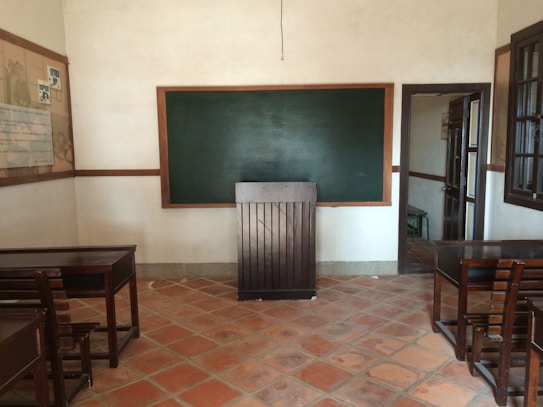 A classroom with a chalkboard on the front wall surrounded by a wooden frame. There are wooden desks and chairs arranged facing the chalkboard. The room has tiled floors and slightly textured beige walls. On the left wall, there is a large board with some documents or posters pinned to it, and on the right, an open doorway leading to another room is visible. A large window is present on the right side of the room.