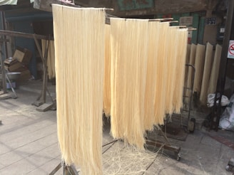Long strands of light-colored noodles are hanging to dry on racks in an outdoor setting. The area appears to be a market or street with some buildings, cartons, and signs in the background. The noodles are suspended from horizontal rods and are evenly spaced, suggesting a process of drying or preparation.