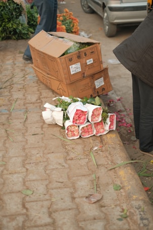Bouquets of pink roses wrapped in white paper are lying on a paved street. Nearby, cardboard boxes are stacked on top of each other with visible labels. Partial views of people and a car can be seen in the background, along with some scattered orange flowers and greenery.