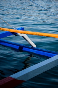 A close-up view of a traditional wooden boat with colorful beams intersecting above the water. The beams are painted in vibrant blue, yellow, and red, contrasting against the rippling surface of the water that reflects the light.