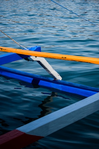 A close-up view of a traditional wooden boat with colorful beams intersecting above the water. The beams are painted in vibrant blue, yellow, and red, contrasting against the rippling surface of the water that reflects the light.