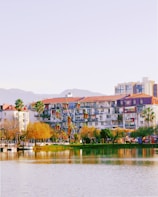 A lakeside view features apartment buildings with balconies and red-tiled roofs, surrounded by palm trees and other greenery. In the foreground, a small Ferris wheel is visible beside the body of water, which reflects the vibrant colors of the scene. Distant hills can be seen under a clear sky.