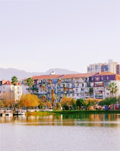 A lakeside view features apartment buildings with balconies and red-tiled roofs, surrounded by palm trees and other greenery. In the foreground, a small Ferris wheel is visible beside the body of water, which reflects the vibrant colors of the scene. Distant hills can be seen under a clear sky.