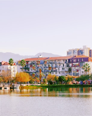 A lakeside view features apartment buildings with balconies and red-tiled roofs, surrounded by palm trees and other greenery. In the foreground, a small Ferris wheel is visible beside the body of water, which reflects the vibrant colors of the scene. Distant hills can be seen under a clear sky.