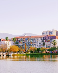 A lakeside view features apartment buildings with balconies and red-tiled roofs, surrounded by palm trees and other greenery. In the foreground, a small Ferris wheel is visible beside the body of water, which reflects the vibrant colors of the scene. Distant hills can be seen under a clear sky.