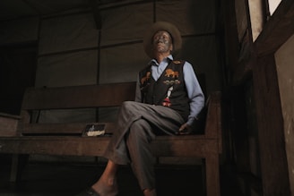 An elderly person sits on a wooden bench in a dimly lit room. They are wearing a wide-brimmed hat, a blue shirt, and a dark vest with colorful animal motifs. The room has a rustic ambiance with simple wooden furniture.