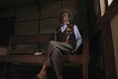An elderly person sits on a wooden bench in a dimly lit room. They are wearing a wide-brimmed hat, a blue shirt, and a dark vest with colorful animal motifs. The room has a rustic ambiance with simple wooden furniture.