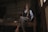 An elderly person sits on a wooden bench in a dimly lit room. They are wearing a wide-brimmed hat, a blue shirt, and a dark vest with colorful animal motifs. The room has a rustic ambiance with simple wooden furniture.