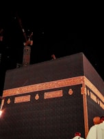 A view of the Kaaba covered in black cloth with intricate golden Arabic calligraphy. A crane is visible in the background against the night sky, and a few people are seen at the bottom corner.