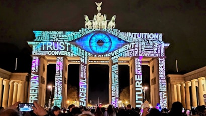 The image depicts the Brandenburg Gate illuminated at night with vibrant projections of words and an eye. The lighting consists of various inspirational words such as 'TRUST', 'RESPECT', and 'PEACEFUL' prominently displayed across the structure, creating a dynamic visual effect. There are people gathered in front, some taking photos.