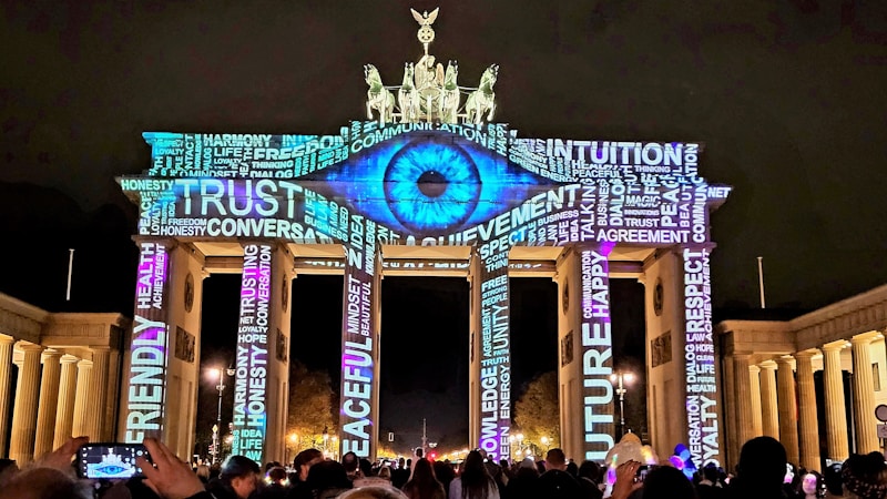 The image depicts the Brandenburg Gate illuminated at night with vibrant projections of words and an eye. The lighting consists of various inspirational words such as 'TRUST', 'RESPECT', and 'PEACEFUL' prominently displayed across the structure, creating a dynamic visual effect. There are people gathered in front, some taking photos.