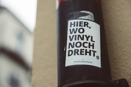A black pole with a white poster attached to it. The poster contains bold black text in German that reads 'HIER, WO VINYL NOCH DREHT'. The background shows a blurred building facade with soft, natural lighting.